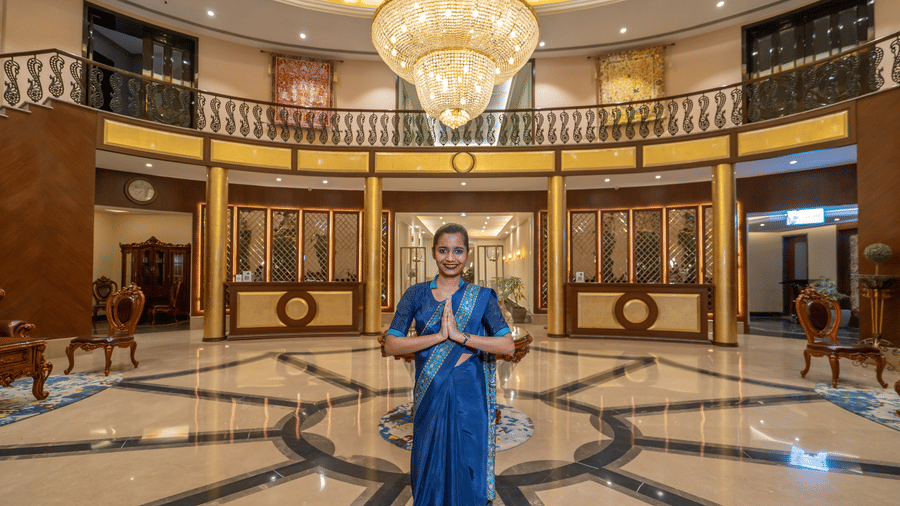 Hotel staff member in a blue uniform standing in the grand lobby, palms together in a traditional Indian greeting at Hotel Hukam's Lalit Mahal.