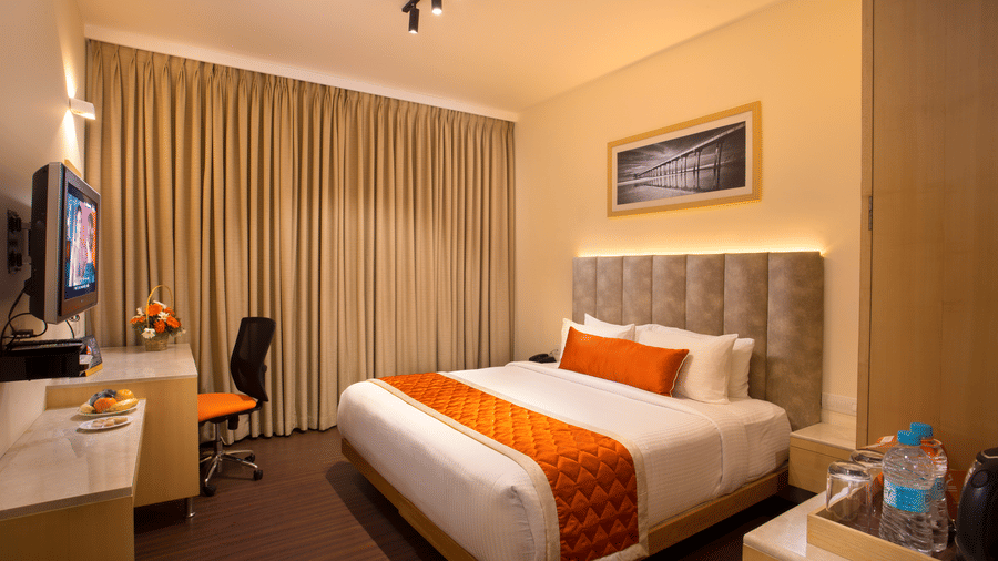 A cosy Premium room at Hotel Langford Keys by TGI featuring a double bed, wall-mounted television, desk with chair, side table with amenities, and large curtained windows.
