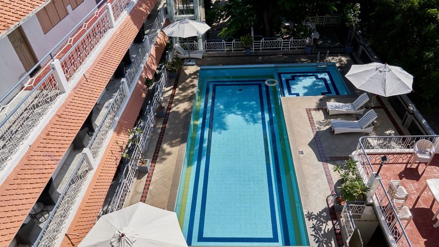A slightly elevated view of a long, rectangular swimming pool at Hotel Mamallaa Heritage, with white umbrellas and the hotel building visible beside it.