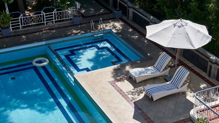 An overhead view of the swimming pool area at Hotel Mamallaa Heritage, showing the blue water, lounge chairs, and white umbrellas on the surrounding deck.