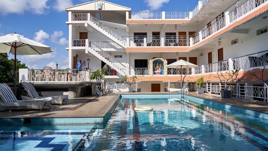 A bright blue outdoor swimming pool at Hotel Mamallaa Heritage, with the multi-storey hotel building visible in the background under a clear sky.