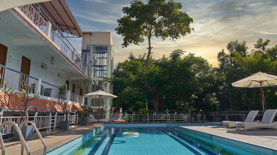 Another view of the refreshing swimming pool area at Hotel Mamallaa Heritage, showing lounge chairs and the white hotel building surrounded by greenery.