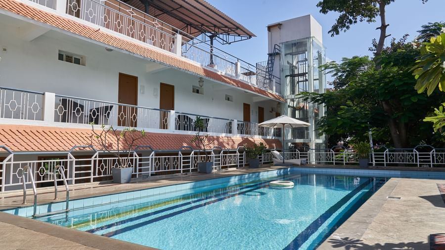 The white multi-storey exterior of Hotel Mamallaa Heritage, with a clear blue swimming pool in the foreground and green trees.