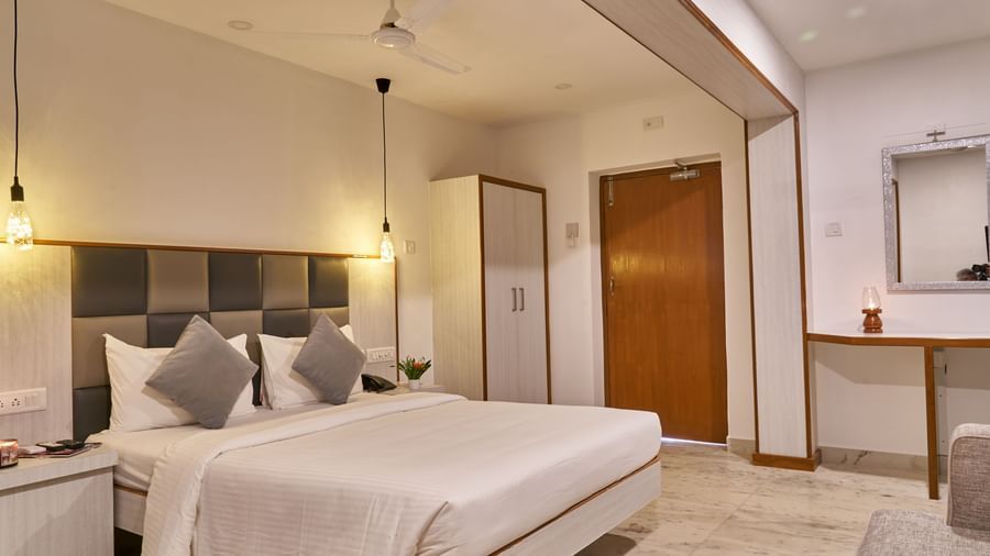 A bright and inviting hotel room at Hotel Mamallaa Heritage, featuring a bed with white linens, a bedside table, and an open doorway leading to another part of the room.