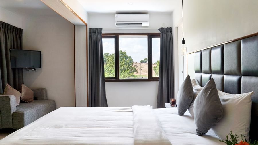 A clean and comfortable hotel room at Hotel Mamallaa Heritage, showcasing a large bed with white linens, a window with a view, and minimalist decor.