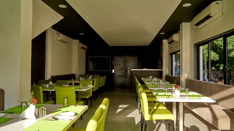 A wide view of a modern dining space at Hotel Mamallaa Heritage, featuring tables with green place settings and a contemporary, bright aesthetic.