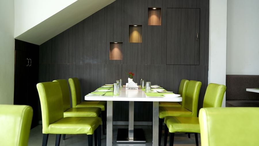 A dining table set with white chairs and vibrant lime green chair backs in a modern restaurant area at Hotel Mamallaa Heritage, illuminated by stylish pendant lights.