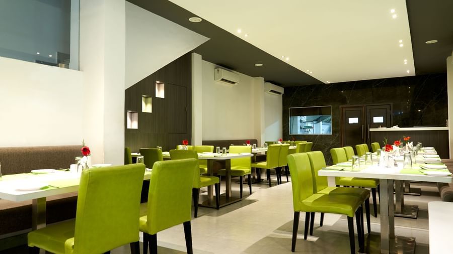 A spacious and contemporary restaurant area at Hotel Mamallaa Heritage, featuring multiple tables set with lime green chairs and white tablecloths.