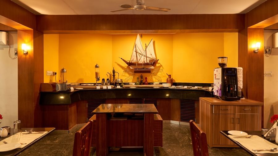 A warmly lit dining area at Hotel Mamallaa Heritage, featuring wooden furniture and a large decorative ship model on a central buffet or counter.