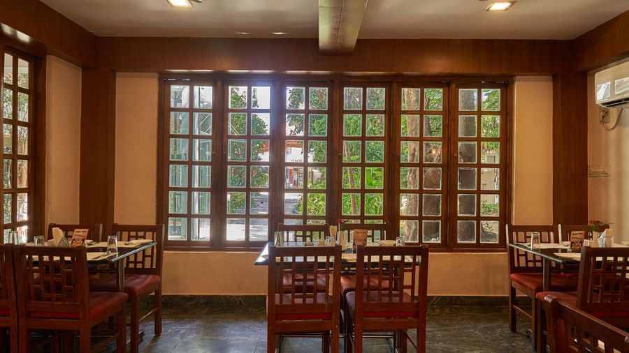 A spacious restaurant interior at Hotel Mamallaa Heritage with numerous large windows framed in dark wood, set with dining tables and chairs.