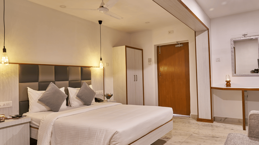 A bright and inviting standard room at Hotel Mamallaa Heritage, featuring a bed with white linens, a bedside table, and an open doorway leading to another part of the room.