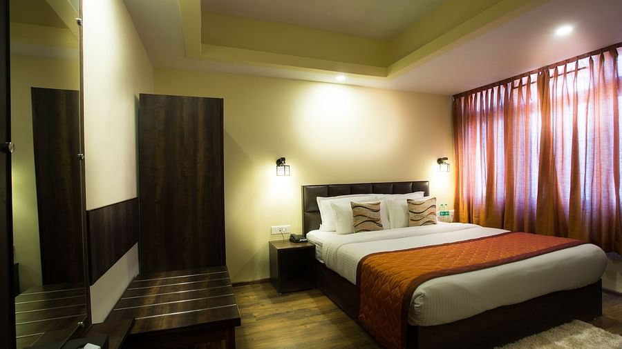 A room at New Orchid Lords Inn Gangtok contains a bed with pillows, bedside tables with lamps and water bottles, curtains, a wardrobe, and a desk.