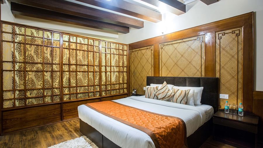 A room at New Orchid Lords Inn Gangtok contains a bed with pillows, a bedside table with water bottles, a rug, wooden accents, and curtains.