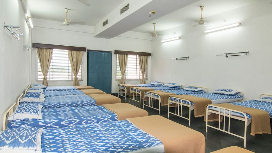 Beds with bedding are arranged in two rows in a room with two windows at Hotel Sahara - Comfortable Dormitory in Ramoji Film City, Hyderabad.