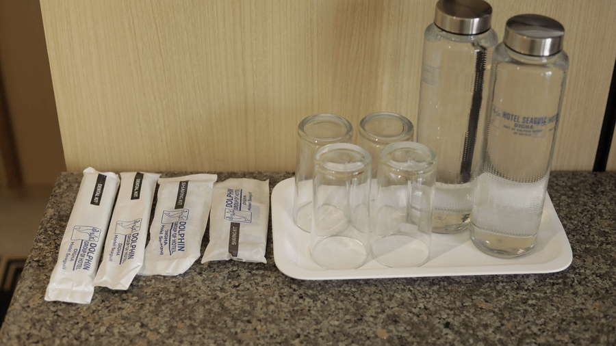A tray holding two glass bottle of water, several clear glasses, and multiple  packets  on a counter set in room at Hotel Seagull Digha