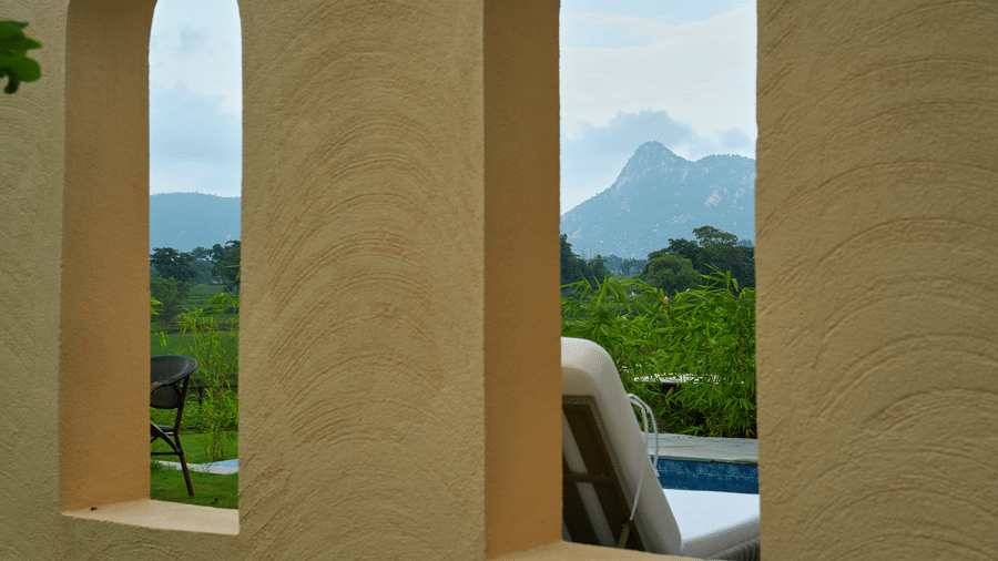 A view through two openings in a wall featuring a lounge chair, green vegetation, and a distant mountain at Hotel Sonar Bangla, Ajodhya Hills.