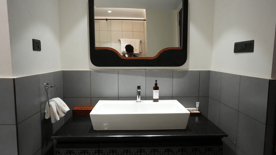 A close-up view featuring a black bathroom vanity, a white basin, and a rounded mirror against grey tiles at Hotel Sonar Bangla, Ajodhya Hills.
