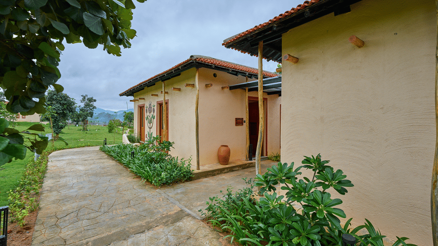 An exterior view featuring a light-coloured building with a terracotta roof next to a paved path and green plants at Hotel Sonar Bangla, Ajodhya Hills.