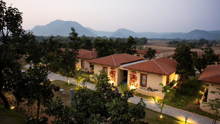 An elevated evening view featuring lit pathways, single-storey cottages with terracotta roofs, and distant hills at Hotel Sonar Bangla, Ajodhya Hills.