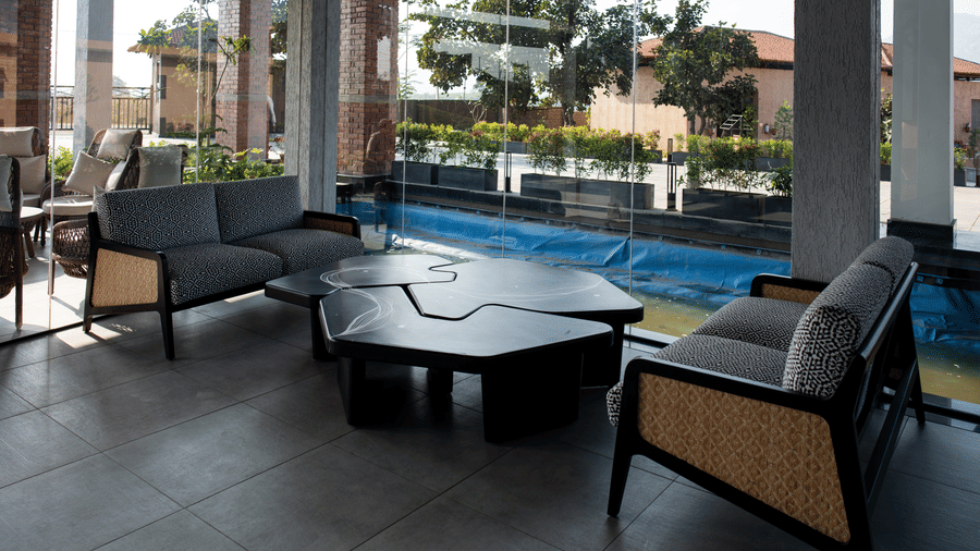 A lounge area featuring two wooden sofas with grey cushions facing an irregular black geometric coffee table beside a glass partition at Hotel Sonar Bangla, Ajodhya Hills.