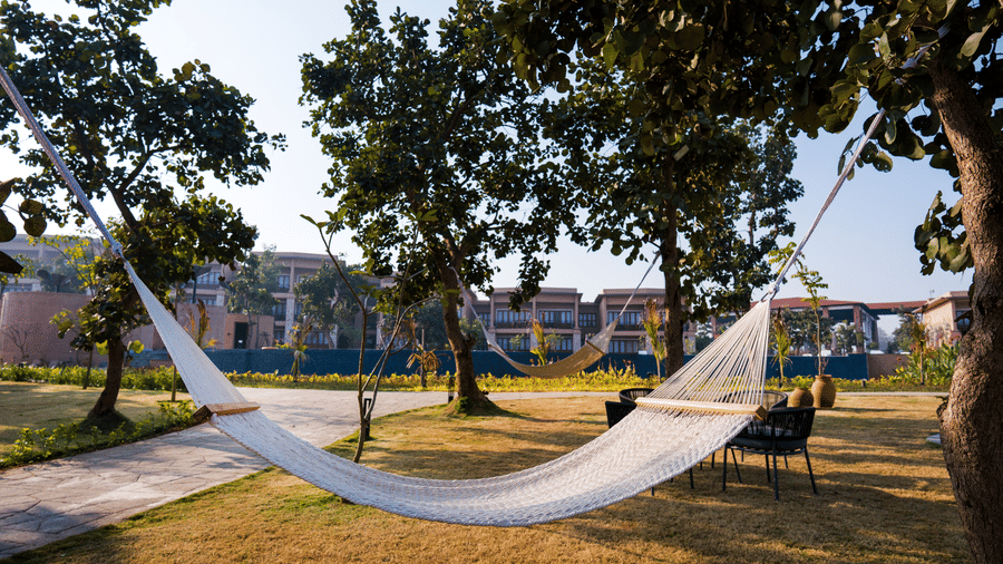 A grassy lawn featuring a white hammock suspended between 2 trees near a paved path at Hotel Sonar Bangla, Ajodhya Hills.
