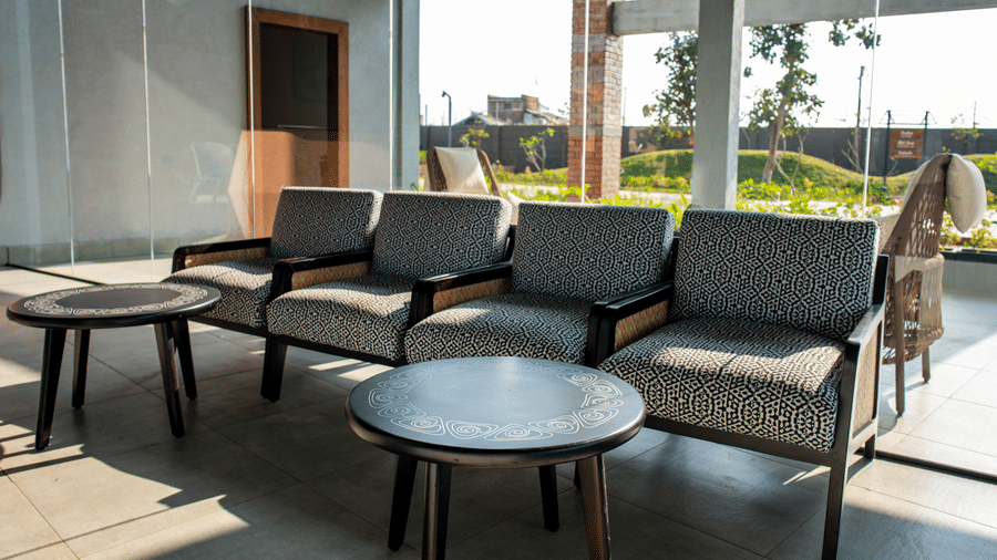 An indoor seating area featuring four grey cushioned armchairs arranged in a row with two round wooden coffee tables next to large floor-to-ceiling windows at Hotel Sonar Bangla, Ajodhya Hills.