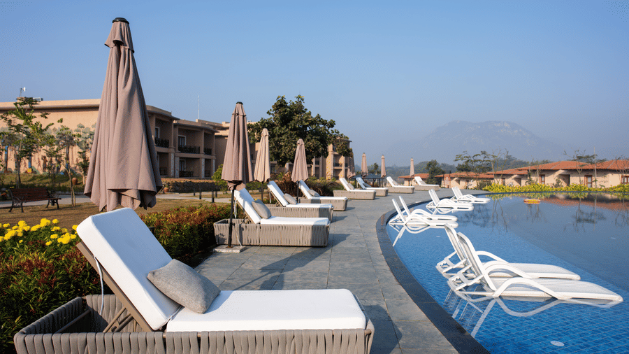 A curved poolside area featuring woven lounge chairs and closed parasols next to the water at Hotel Sonar Bangla, Ajodhya Hills.