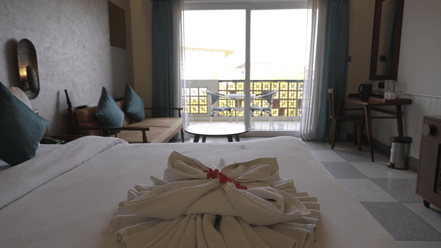 A guest room showing a double bed with towel art in the foreground and a seating area by the balcony at Hotel Sonar Bangla, Mayapur.