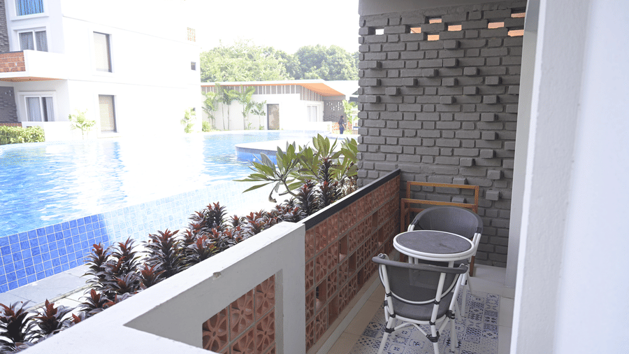 A balcony with a grey chair and table overlooking a blue swimming pool and hotel buildings at Hotel Sonar Bangla, Mayapur.