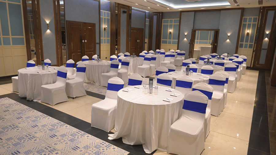 Round banquet tables arranged with white covers and blue chair sashes in a spacious event hall at Hotel Sonar Bangla, Mayapur.