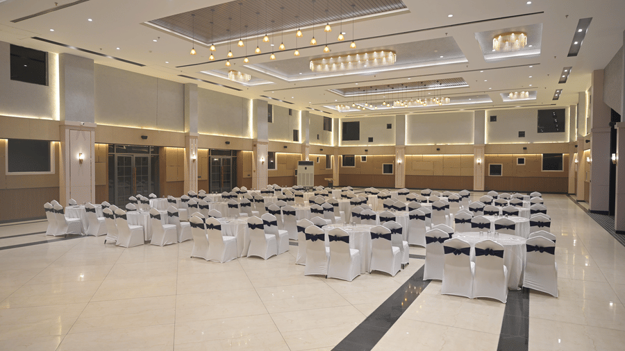 A panoramic view of the banquet hall arranged for a gathering, featuring bright chandeliers and neatly placed round tables at Hotel Sonar Bangla, Mayapur.