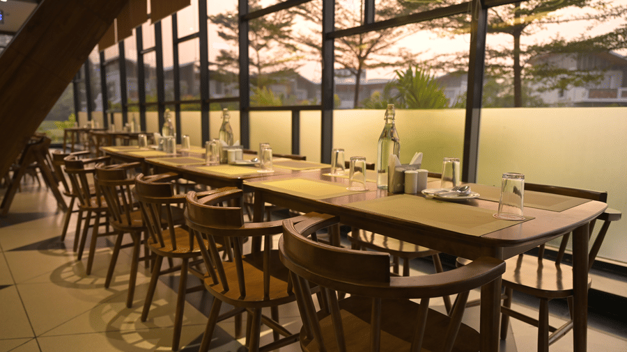 A long wooden dining table set for several people next to floor-to-ceiling windows at Hotel Sonar Bangla, Mayapur.