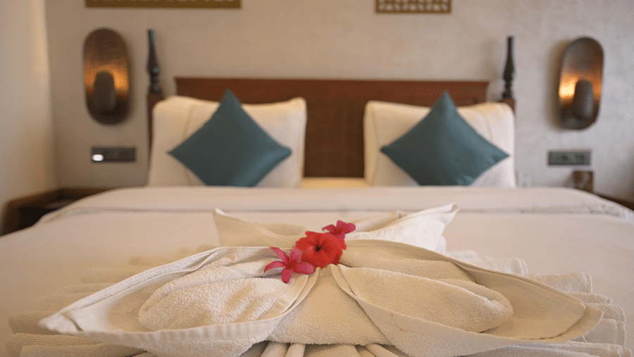 A double bed with white linen, teal cushions, and a pink flower on a decorative towel arrangement at Hotel Sonar Bangla, Mayapur.