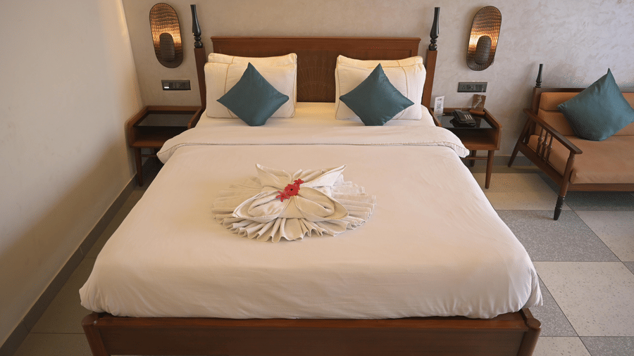 A front-facing view of a double bed with white bedding, blue cushions, and a decorative flower on a towel at Hotel Sonar Bangla, Mayapur.