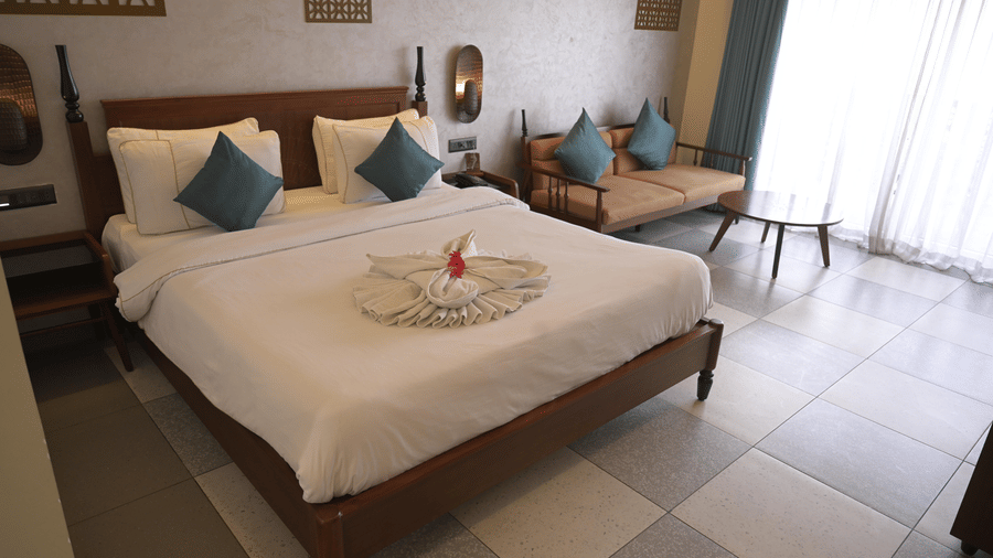 A guest room with a double bed, white linens, and blue pillows with a wooden sofa in the background at Hotel Sonar Bangla, Mayapur.