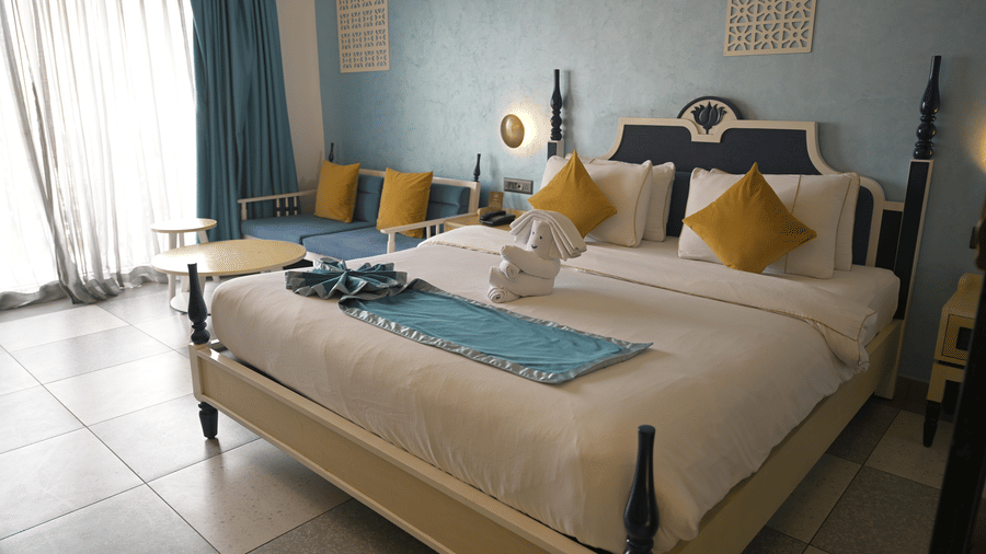 A wide shot of a guest room featuring a bed with white and blue linens and yellow pillows at Hotel Sonar Bangla, Mayapur.