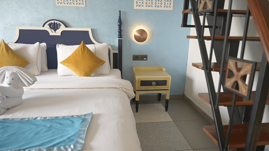 A bed with white linens, yellow pillows, and a blue runner next to a wooden staircase at Hotel Sonar Bangla, Mayapur.