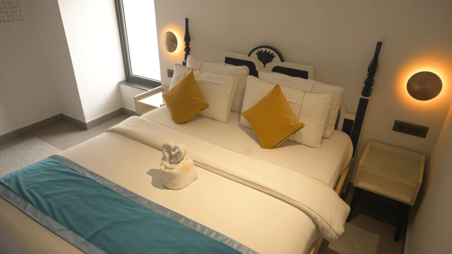A double bed with white linens, yellow cushions, and a blue bed runner flanked by circular wall lamps at Hotel Sonar Bangla, Mayapur.