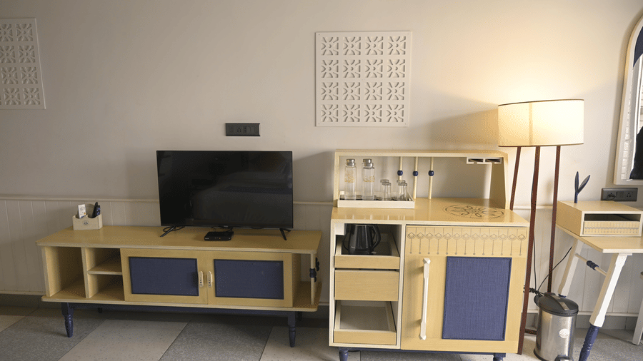  A television on a blue and yellow console table next to a refreshment cabinet and a floor lamp at Hotel Sonar Bangla, Mayapur.