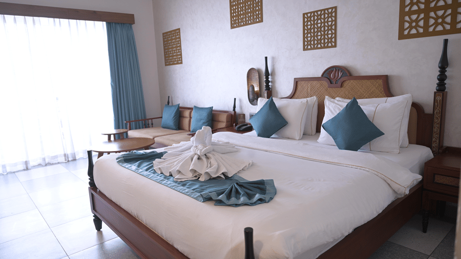 A guest room featuring a double bed with towel art, wall decorations, and a window with blue curtains at Hotel Sonar Bangla, Mayapur.