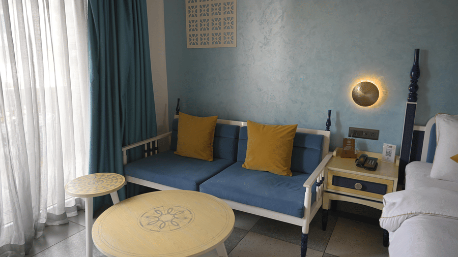 A blue sofa with yellow throw pillows next to a window with blue curtains at Hotel Sonar Bangla, Mayapur.