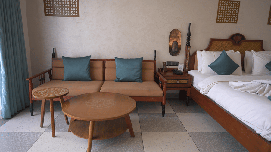 A wooden sofa with blue cushions and 2 round wooden coffee tables in a guest room at Hotel Sonar Bangla, Mayapur.