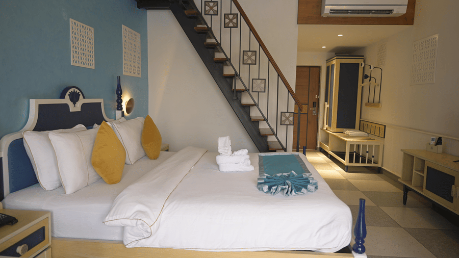 A guest room bed positioned next to a wooden staircase with metal railings at Hotel Sonar Bangla, Mayapur.