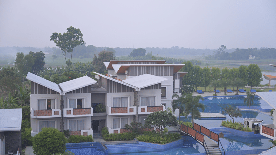 An outdoor view of white multi-storey hotel buildings and a swimming pool with a wooden bridge at Hotel Sonar Bangla, Mayapur.