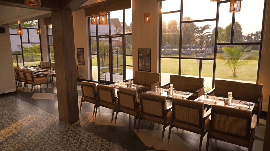 A sun-lit dining space with wooden tables, cushioned chairs, and expansive windows overlooking the lawn at Hotel Sonar Bangla ,Mayapur.