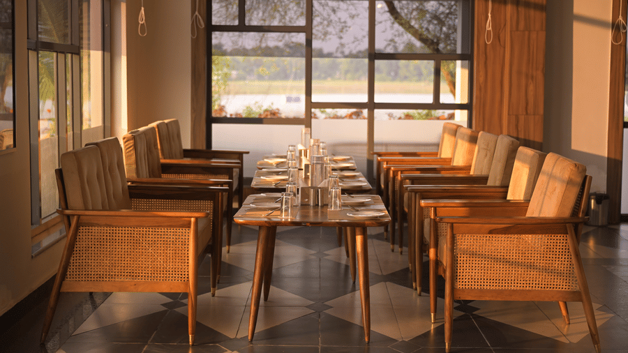 An indoor seating area with wooden chairs and large windows framing outdoor garden views at Hotel Sonar Bangla, Mayapur.