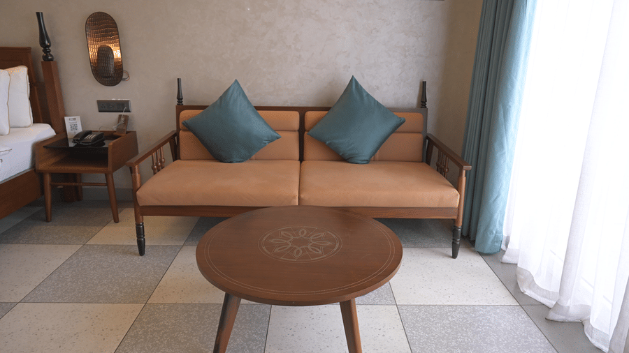 A wooden sofa with teal cushions and a round wooden coffee table next to a window at Hotel Sonar Bangla, Mayapur.
