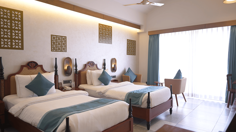  A wide shot of a twin bedroom featuring wooden headboards, blue accents, and a ceiling fan at Hotel Sonar Bangla, Mayapur.