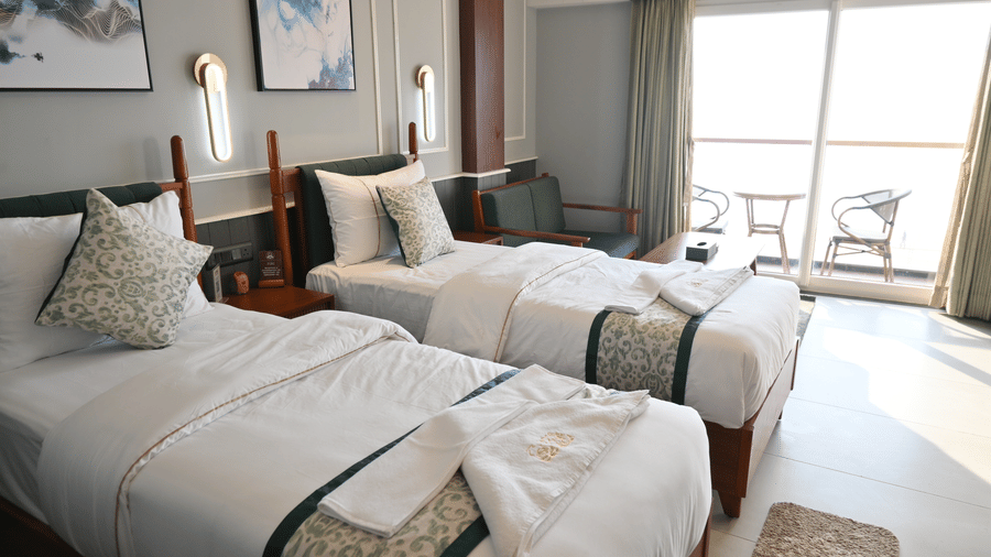 A modern hotel twin room featuring two single beds, dark wooden furniture, and a sliding glass door opening onto a bright balcony with a view at Hotel Sonar Bangla, Puri.