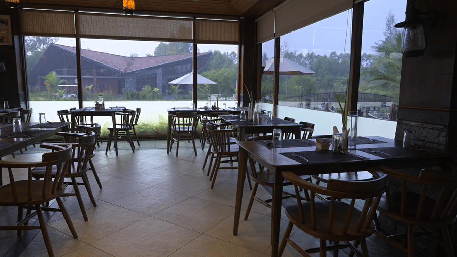 All-day dining area at Hotel Sonar Bangla, Sundarban with wooden tables, spindle-back chairs, large glass walls, and a view of the property exterior.